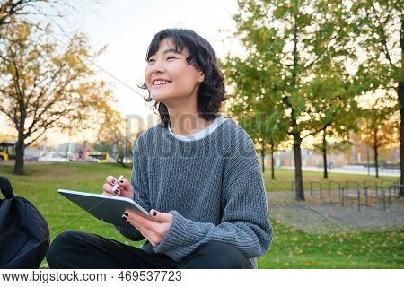 Young Korean Woman Sits In Park, Draws On Digital Tablet, Writes In Gadget With Pen Tool, Looks Arou