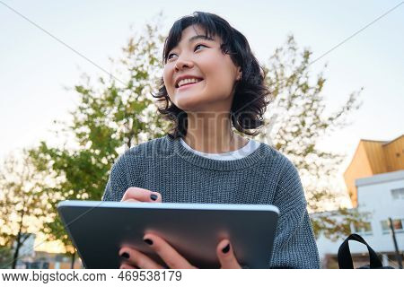 Close Up Portrait Of Young Korean Girl Sits Outdoors In Park, Holds Her Digital Tablet And Graphic P