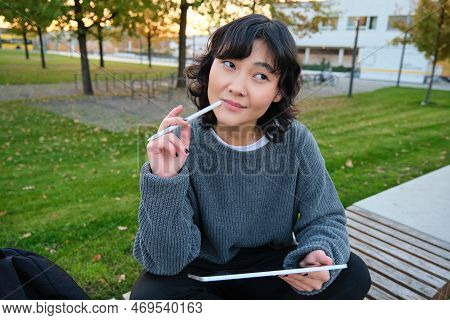 Portrait Of Asian Woman, Student In Park, Sits On Bench With Digital Tablet And A Pen, Thinking, Loo