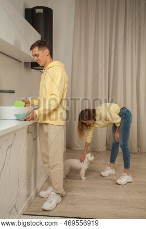 Happy Cherrful Couple In The Kitchen In Yellow Hoodies, Man Washing The Dishes And Woman Petting Lit