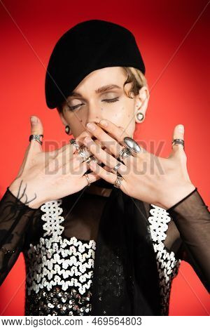 Queer Person With Closed Eyes Covering Mouth With Hands In Silver Rings On Red Background.