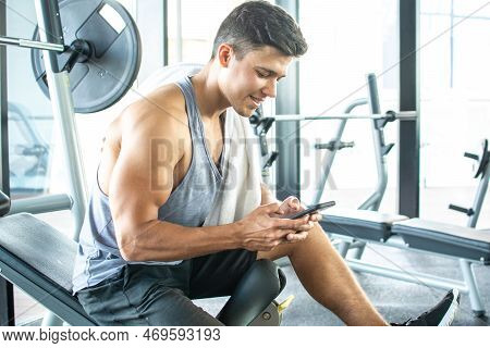 Handsome Young Man Resting On Weight Bench And Using Phone After Training At Gym.