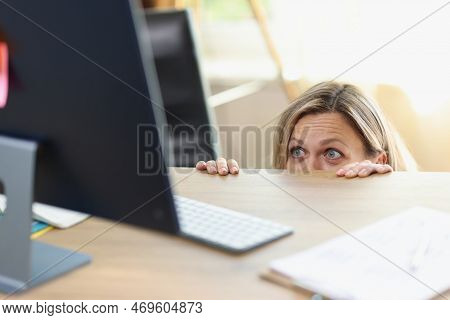 Scared Female Manager Hidden Behind Office Desk And Looking At Computer Screen. Unwanted Communicati