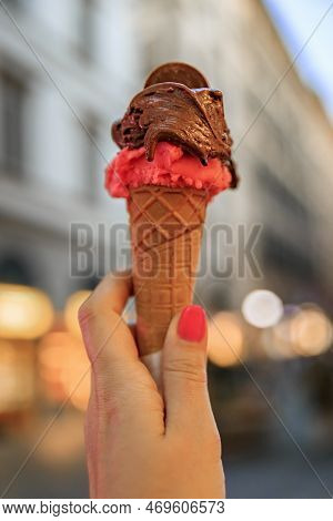 Hand Holding An Artisanal Chocolate And Raspberry Gelato, A View Of Blurred Houses And Lights On A W