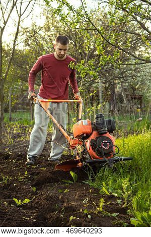 A Farmer Man Works In The Field, Plowing The Land With A Plow On The Farm. A Plowman On A Walk Behin