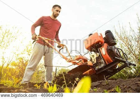 A Farmer Man Works In The Field, Plowing The Land With A Plow On The Farm. A Plowman On A Walk Behin