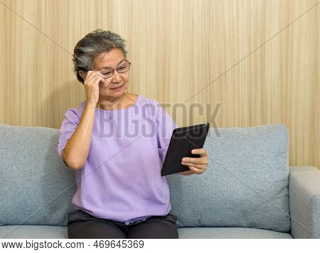 Asian Retired Elderly With Eyeglasses, Sitting On The Sofa Watching At Tablet Computer. With Presbyo