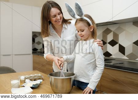 Mother And Her Preschool Daughter In Bunny Ears Preparing And Whisking Dough In Modern Kitchen. Smil