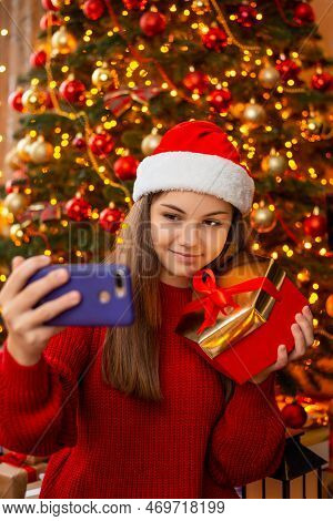 Cheerful Young Girl In Santa Hat, Making Selfie With Gift Box Near Christmas Tree. Winter Holidays H