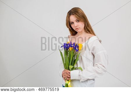 A Blonde Girl In A White Shirt And Blue Jeans Holds A Bouquet Of Tulips And Irises Sitting On A Whit