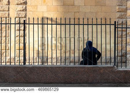 Back View Of Unrecognizable Arab Traditional Woman With Hijab On, Sitting On The Edge Of A Curbstone