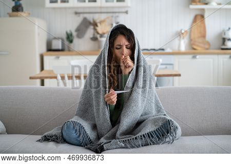 Frightened Unhealthy Woman Looking At Thermometer Sits Wrapped In Warm Plaid On Sofa In Home Interio