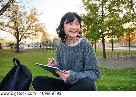 Young Korean Woman Sits In Park, Draws On Digital Tablet, Writes In Gadget With Pen Tool, Looks Arou
