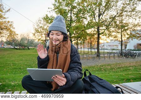 Portrait Of Young Asian Girl Sits In Warm Clothes In Park, Waves Hand At Tablet, Video Chats Outdoor