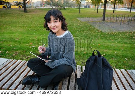 Young Asian Girl With Graphic Pencil And Tablet, Sits In Park On Bench, Draws Scatches, Does Her Hom