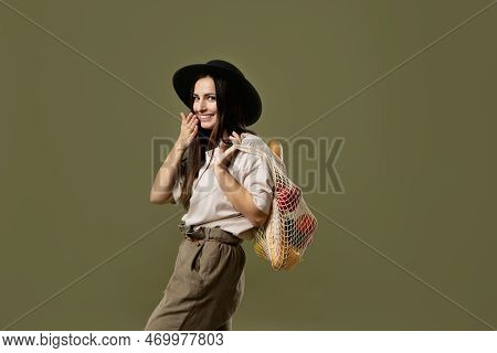 Cherfull Brunette Woman In Beige T-shirt And Black Hat Holding A Mesh Eco Bag Full Of Fruets And Veg
