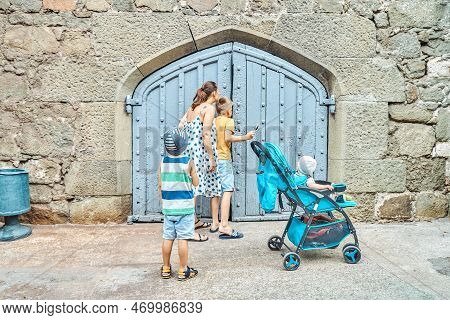 Family Members Walk Together And Try To Look Through Wooden Gate In Stone Wall. Tourists Spend Vacat
