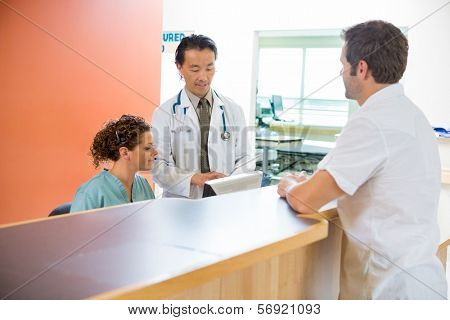 Male doctor and nurse using digital tablet while patient standing at reception desk in hospital