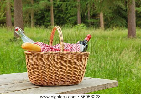 A whicker picnic basket full of food and drink on a table in a woodland setting on a bright summers day.