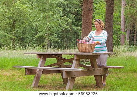 A woman placing a whicker picnic basket full of food and drink on a table in woodland, on a bright summers day.