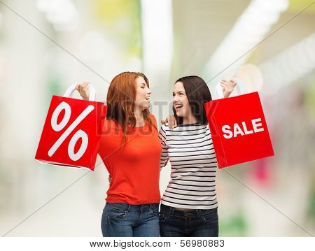 shopping, sale, mall and gift sconcept - two smiling teenage girls with shopping bags and percent sign at shopping mall