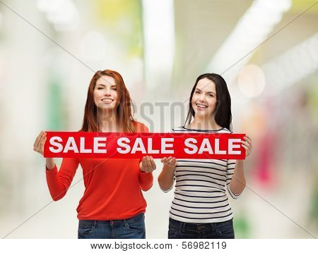 shopping, sale, mall and gift sconcept - two smiling teenage girls with sale sign at shopping mall