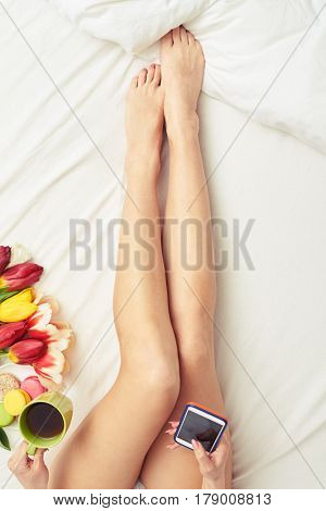 Overhead shot of slim long female hands isolated on the bed. Cropped shot of female lying on the bed. Drinking tea. White bed. Flowers lying on the white surface 
