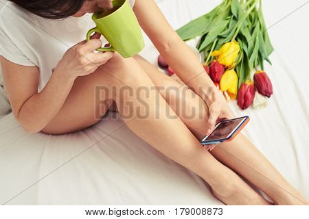 Overhead shot of bared female legs sitting on the bed. Woman sipping tea from the cup. Flowers lying next to the woman. Perfect morning in the bed