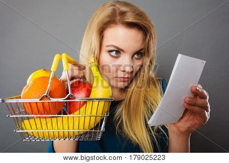 Worried woman holding shopping basket with fruits looking at bill receipt being scared of huge prices