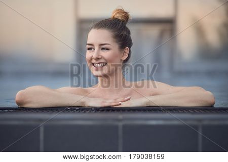 Happy young woman enjoying a relaxing swim standing in a swimming pool resting her hands on the tiled surround grinning as she looks to the side