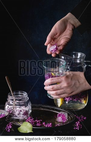 Femail hands put ice cubes with flowers into glass of lilac lemonade with lemon. Glass jar of sugared lilac flowers and pitcher on black tablecloth over black. Dark rustic atmosphere. See series.