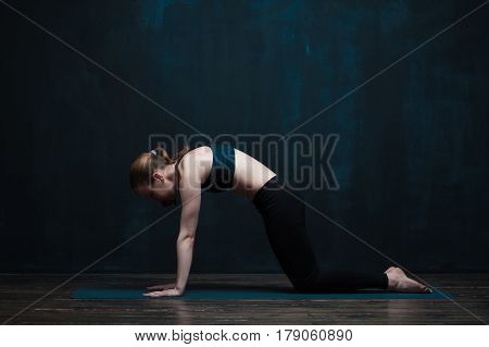 Sporty young woman doing body balance exercises on blue mat. Fit girl wearing black sportswear practicing yoga asana against dark wall. Fitness and healthy lifestyle concept.