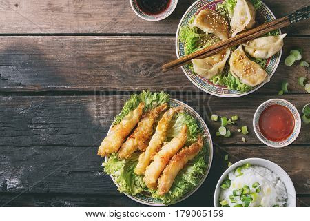 Fried tempura shrimps and gyozas potstickers on lettuce salad with sauces and rice. Served in traditional china plate with chopsticks over old wooden background. Top view. Asian dinner