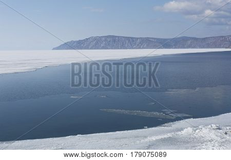 Baikal lake spring landscape view. Snow-covered shore of the lake. Rocky forested coastline. Boundary of ice and open water