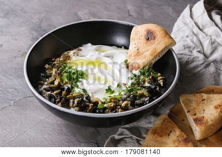 labneh middle eastern lebanese cream cheese dip with olive oil, salt, herbs, olives tapenade served in black bowl with traditional pita bread over gray texture metal background.
