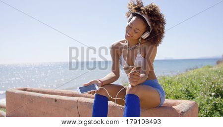 Smiling female wearing swimsuit top and shorts sitting on crb listening music with headphones on ocean background.
