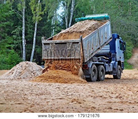 Blue dump truck with hoisted body unloads sand on the building site in rural areas