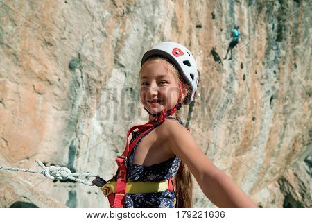 Little Rock Climber in protection climbing Helmet and safety Harness taking self Portrait Picture with high vertical outdoor Wall and other Athletes on Background