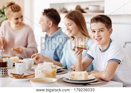 So delicious. Cheerful contewnt boy sitting at the table and eating tasty food while having breakfast