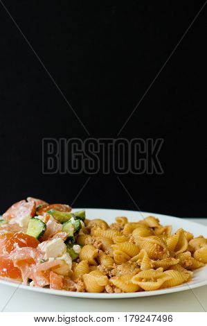 Vertical photo of italian pasta conchiglie with chicken mince sauce and fresh vegetable salad with feta cheese on white plate black background with space for text
