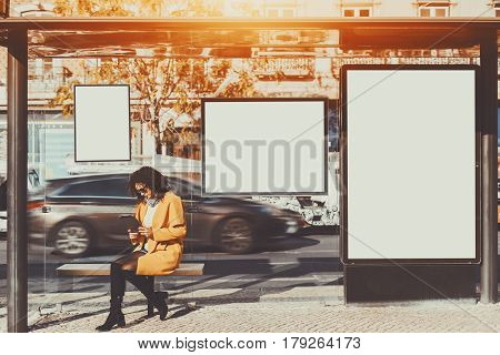 Curly brunette woman in sunglasses and yellow coat working on her digital tablet while sitting and waiting bus inside of glass city bus stop with several blank mock-up banners around her