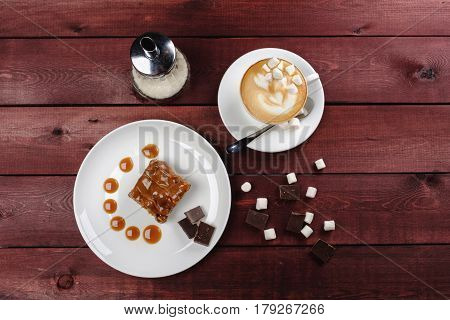 a piece of tasty chocolate brownie and caramel sauce a cup of cappuccino and marshmallows on a white plate on a wooden table. Top view