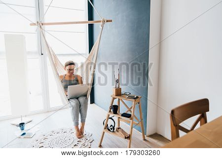Young freelancer girl sitting in a wicker hammock at home with laptop. A female blogger works at home. Work on the Internet.