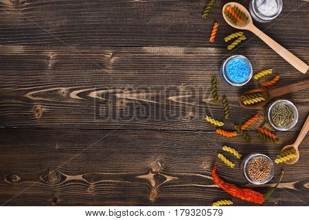 Dried fusilli colorful pasta salt white and blue spices herbs in glass jars red chilly peppers and spoons on brown wooden background. Ingredients for cooking and seasoning food copy space