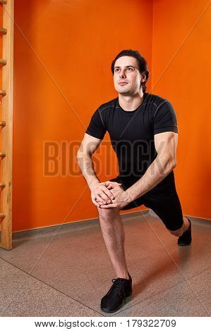 beautifull strong athletic guy, execute exercise in sport-hall in the gym. Sportsman in the black sportwear, t-shirt and shorts, black sneakers. Background of the bright orange wall. Squatting. Knee. Healthy lifestyle.