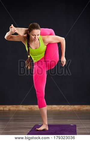 Beautiful young woman working out indoors, doing yoga exercise on violet mat, standing in Vrksasana Posture, Tree Pose. Woman in the sportwear, pink leggings and green t-shirt. Healthy lifestyle.