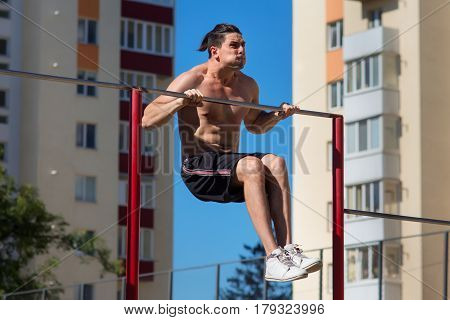 Fit young man doing push ups on horizontal bar outdoors with big effort. Man in the black short and white sneakers. Abs. Haelthy lifestyle. Park. Outside. Spring. Face and emotions.