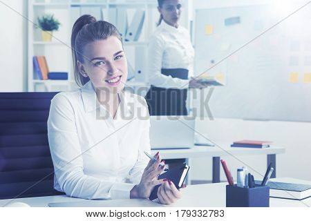 Portrait of a smiling blond businesswoman with notebook at her workplace in a white office. Her colleague is in the background making a drawing on a whiteboard. Toned image