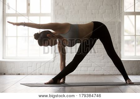 Young yogi woman practicing yoga concept, standing in Utthita Trikonasana exercise, extended triangle pose, working out, wearing sportswear, full length, white loft studio background, side view