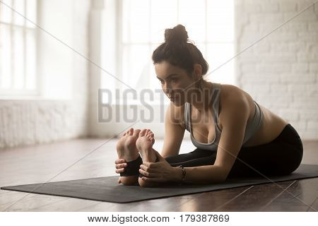 Young attractive yogi woman practicing yoga concept, sitting in paschimottanasana exercise, Seated forward bend pose, working out on black mat, wearing black sportswear, white loft studio background